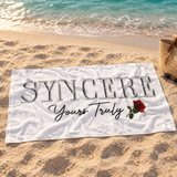 White SYNCERE Yours Truly rose beach towel spread on sand beside turquoise water